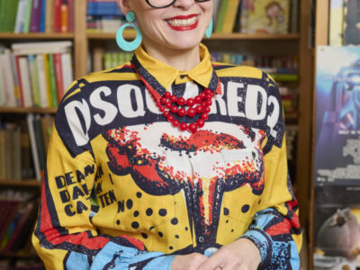 Frau mit Brille, roter Kette und gelbem Dsquared Hemd vor Bücherregal. Mode, Stil, Accessoires.
