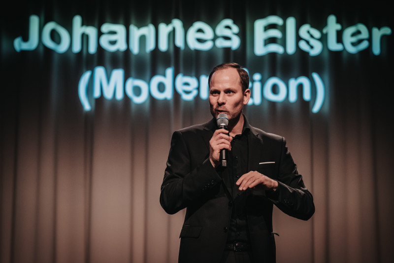 Johannes Elster, Moderator auf der Bühne mit Mikrofon. Event-Moderation, Bühnenauftritt.