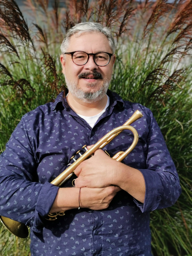 Mann mit Brille hält Trompete vor grünem Hintergrund. Musikerporträt, Instrument, Musikveranstaltung.