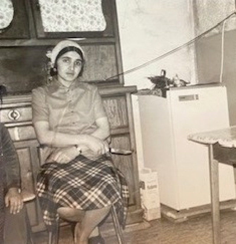 Historisches Foto: Frau sitzt in Küche, trägt Kopftuch und karierten Rock. Vintage-Interieur mit Kühlschrank. Nostalgie pur!